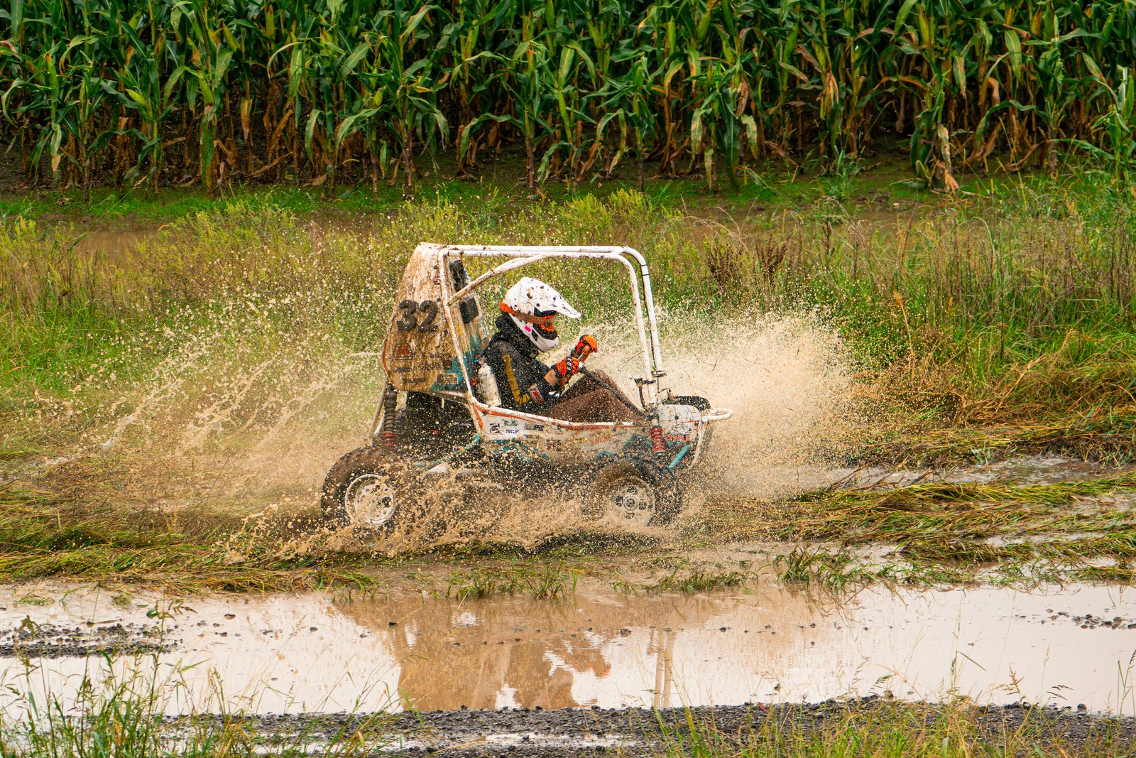 Baja car number 32 blasting through a muddy water hole in front of a cornfield, spray rising around the car