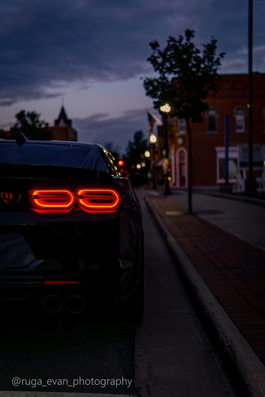 Lit Camaro taillights in the foreground with a downtown street receding at dusk, vertical frame