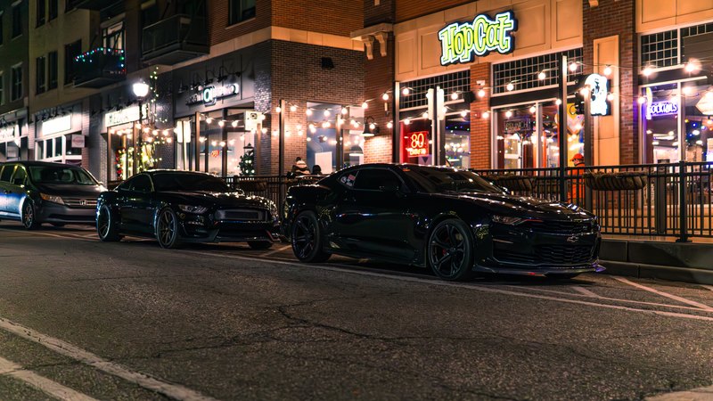 A black Mustang and a black Camaro parked together on a lit downtown street at night