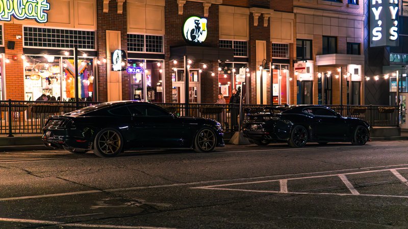 Black Mustang and Camaro on a lit downtown street at night