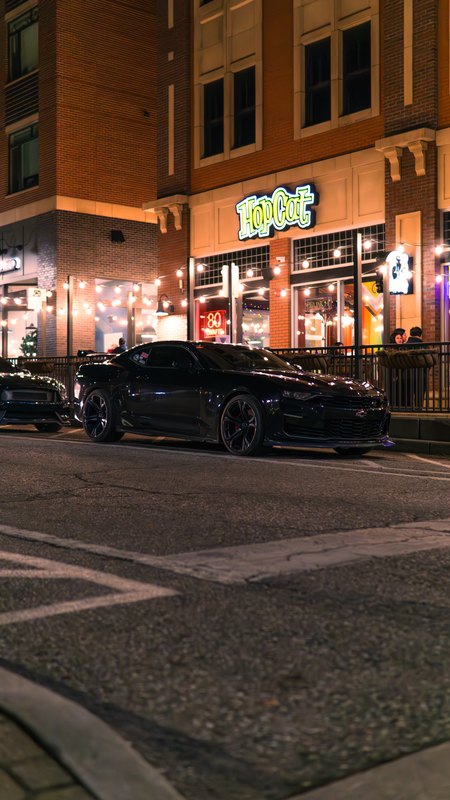 Black Mustang and Camaro on a lit downtown street at night